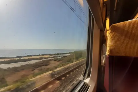 Blick aus dem Zugfenster auf eine Küstenlandschaft mit Meer und Bahngleisen. Die Reise mit dem Zug nach Mallorca beginnt.