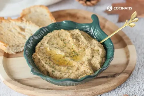Delicioso pate de berenjena cremoso, servido en un cuenco verde con un toque de aceite de oliva y perejil. Acompañado de tostadas crujientes.