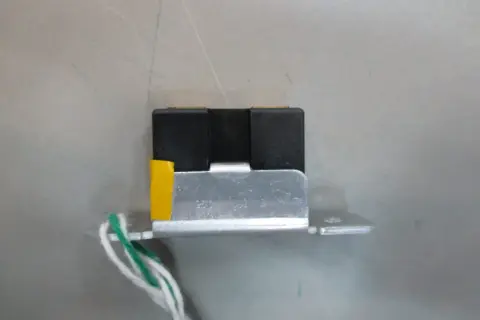 Mały, czarny element z żółtym akcentem i przewodami, przypominający wtyczkę RGB, zamontowany na metalowej płytce.