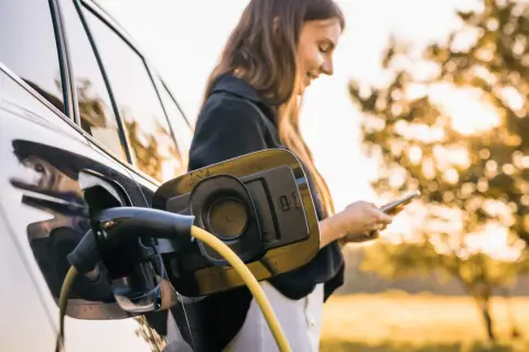En kvinna laddar sin elbil utomhus, med solen som lyser genom träden. En gul laddningskabel är ansluten till bilen.