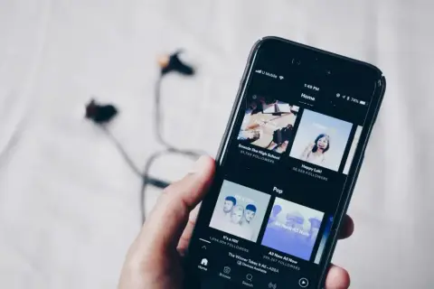 Dłoń trzyma smartfon z aplikacją do streaming muzyki. W tle leżą słuchawki.