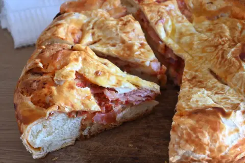 Una torta rustica con pasta sfoglia ripiena di salumi e pomodoro, tagliata a fette su un tagliere di legno.