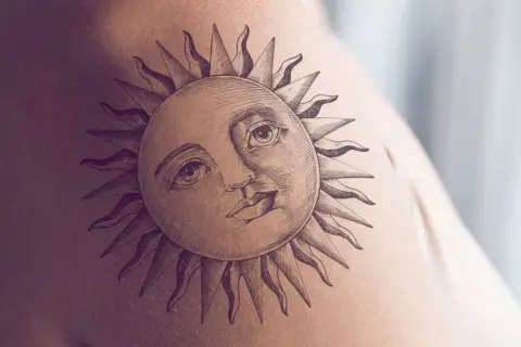 Ein detailreiches Tattoo einer Sonne mit menschlichem Gesicht und Strahlen auf der Haut. Dieses sonnenstrahlen tattoo strahlt Wärme aus.