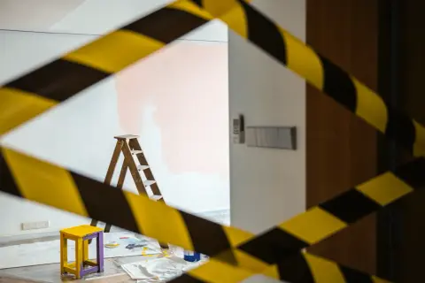 Cinta de precaución amarilla y negra rodea una habitación con una escalera, un taburete y pintura rosa en la pared, ilustrando la diferencia entre obras de mejora y obras de conservación.
