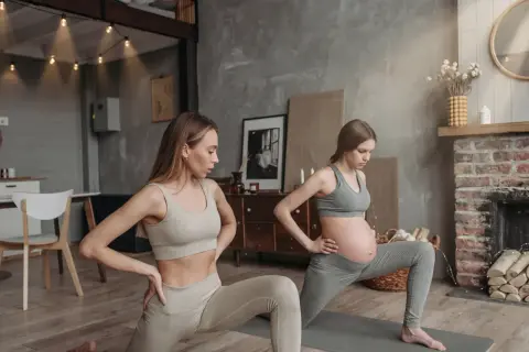 Två kvinnor gör yoga gravid i ett mysigt rum. Den ena kvinnan är gravid och den andra är hennes träningskompis.