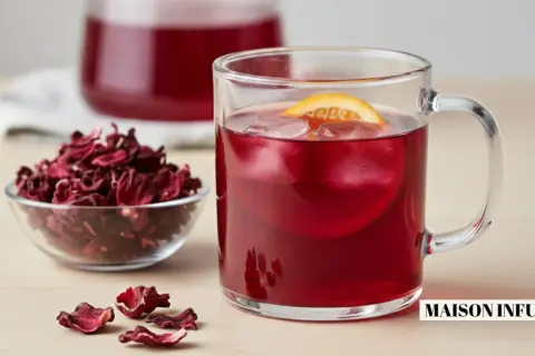 Verre de boisson rouge vif avec glaçons et tranche d'orange, à côté d'un bol de fleurs d'hibiscus séchées. L'eau d'hibiscus, rafraîchissante et colorée.
