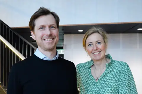 Tobias Sahlin ler glatt bredvid en kvinna i grön mönstrad blus. De står framför en trappa med belysning.
