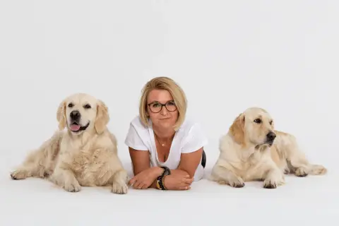 Kobieta w okularach leży między dwoma golden retrieverami na białym tle.