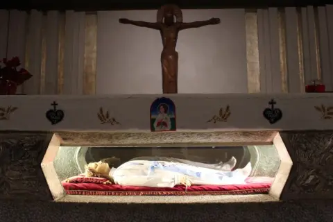 Relikwie najmłodszego świętego spoczywają w szklanej gablocie. Nad nimi drewniany krzyż i wizerunek Matki Bożej.