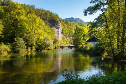 Rivière paisible reflétant la verdure luxuriante, un pont discret et une maisonnette au bord de l'eau, le tout sous un ciel bleu clair, évoquant la sérénité de la cascade de Syratus.