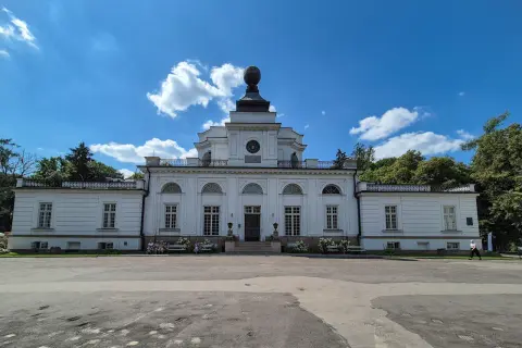Pałac w Jabłonnie, biała, klasycystyczna budowla z charakterystyczną kopułą, otoczona zielenią, pod błękitnym niebem z chmurami.