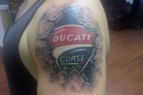 Tatuaje de un logo de Ducati Corse en el hombro, con efecto de piel rasgada.