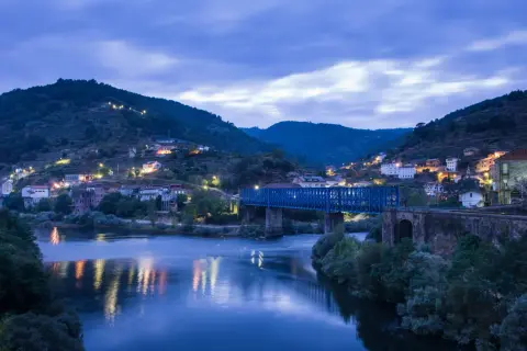 Un puente azul cruza un río tranquilo, reflejando las luces de un pueblo en la ladera. La noche cae sobre este paisaje, creando una atmósfera serena.