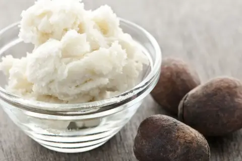 Schüssel mit cremiger Parkii Butter neben drei braunen Nüssen auf Holzhintergrund.