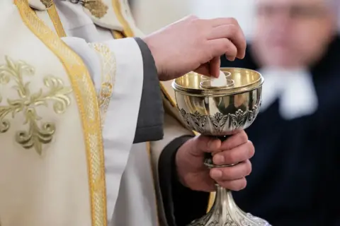 Dłoń księdza w białej szacie z haftem podaje hostię do kielicha. Komunia bez spowiedzi, symbol wiary.