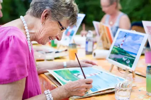 Des personnes âgées participent à des activités manuelles en peignant en plein air. Une dame souriante, vêtue d'un haut rose, est concentrée sur sa toile.