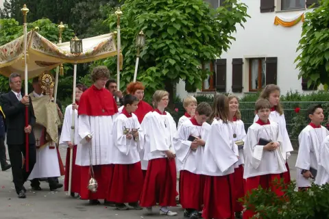 Prozession am Corpus Christi Feiertag mit Ministranten in weißen und roten Gewändern, die eine Monstranz tragen.