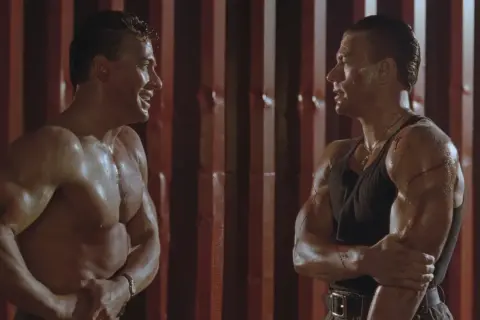 Dwaj Jean-Claude Van Damme w scenie z jednego z jego filmów, spoceni i z widocznymi ranami, patrzą na siebie.