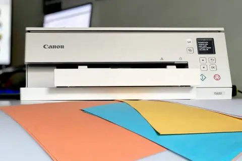 Drukarka Canon TS6351 wyświetla komunikat o błędzie, sugerując, dlaczego z drukarki wychodzą puste kartki. Na biurku kolorowe papiery.