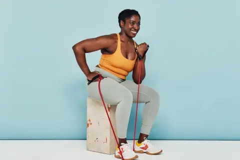 Woman smiles while performing arm strength exercises with a resistance band, seated on a box.