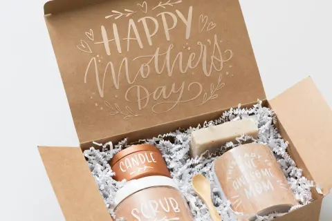 Otwarty brązowy karton z napisem "Happy Mother's Day" i zawartością: świeca, peeling, mydło, kubek i drewniana łyżeczka.