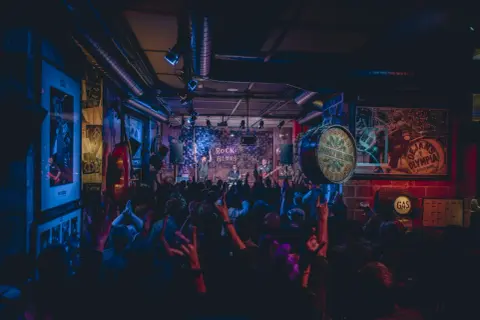 Una banda toca rock y blues en un local con luces de neón. El público levanta las manos, creando una atmósfera vibrante, como si estuvieran en una gran casa en Zaragoza.