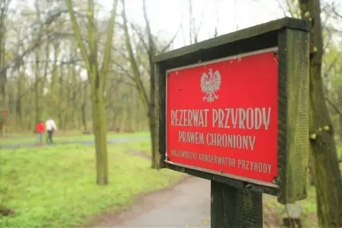 Czerwony znak informuje o rezerwacie przyrody. W tle spacerują dwie osoby pośród drzew.