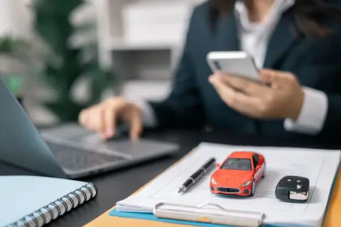 Biznesmen analizuje amortyzację samochodu osobowego 75 czy 100, korzystając z telefonu i laptopa. Na biurku model auta i kluczyki.