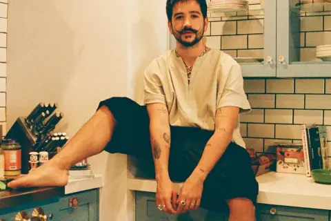 Camilo Alonso Vega, con su característico bigote, posa relajado en una cocina. Sus tatuajes y anillos añaden un toque personal a su estilo.