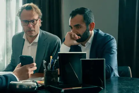 Två män i kostym sitter vid ett bord. Den ena mannen med glasögon tittar på sin mobiltelefon, den andra mannen funderar. Scen från "Hundarna" på SVT.