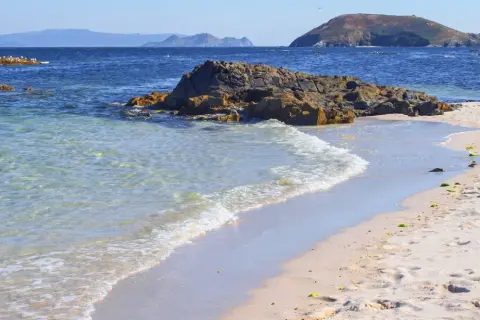 Aguas cristalinas acarician las **islas de Ons playas** de arena dorada. Rocas oscuras emergen del mar azul, mientras gaviotas planean en el cielo despejado.