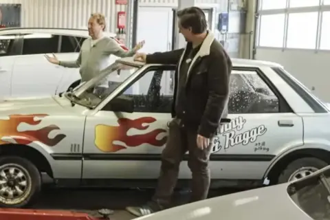 Två män vid en silverfärgad bil med flammor och texten "Ronny & Ragge".