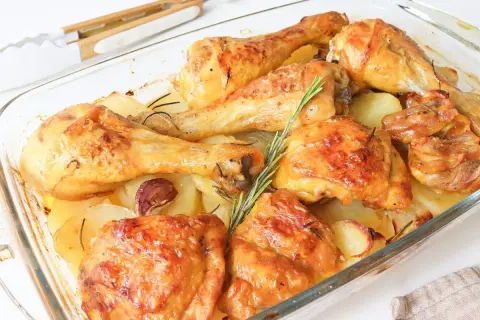 Muslos de pollo al horno con patatas, dorados y jugosos, acompañados de patatas asadas, ajo y romero en una fuente de cristal.