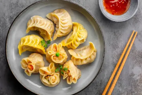Talerz pełen różnorodnych pierożków dim sum, z których każdy wygląda jak małe dzieło sztuki. Obok miseczka z sosem i pałeczki.