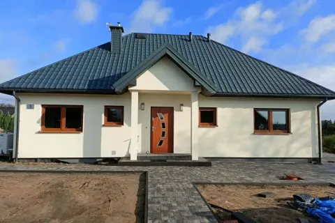 Nowoczesny dom kanadyjski z jasną elewacją, ciemnym dachem i drewnianymi oknami. Przed domem ścieżka z kostki brukowej i plac budowy.