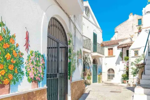 Vicolo fiorito nel centro storico di Rodi Garganico, con murales colorati su muri bianchi e un cielo azzurro brillante.