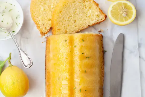 Un delizioso plumcake al limone glassato, con fette affettate, limoni freschi, una ciotolina di glassa e un coltello.