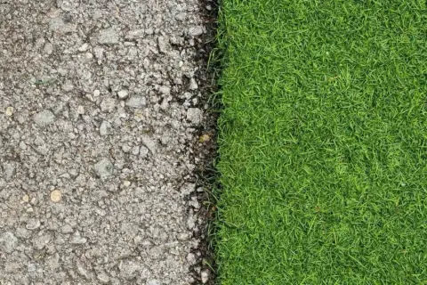 Superficie de asfalto gris y césped natural verde vibrante, mostrando la transición de poner césped artificial sobre césped natural.