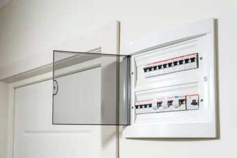 Otwarta biała skrzynka z bezpiecznikami elektrycznymi i przezroczystą klapką.