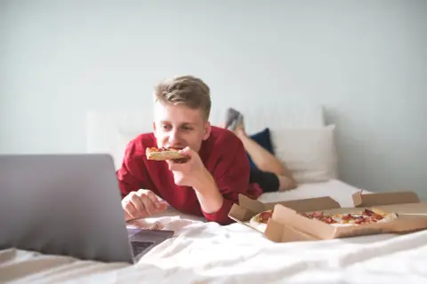 A young man eats pizza in bed while watching a laptop. Is it bad to go to sleep after eating? This scene suggests a late-night snack before bed.