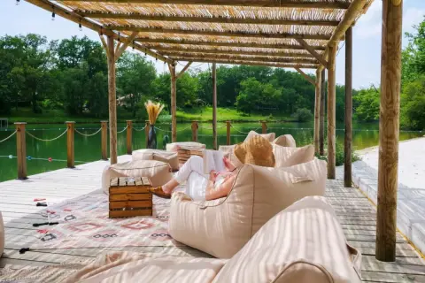 Détente sous le toit de chaume du whaka lodge, au bord d'un lac paisible. Une femme lit, entourée de poufs confortables.