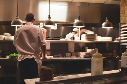Un ayudante de cocina en España prepara ingredientes frescos en una cocina profesional, con pilas de platos y utensilios listos para el servicio.