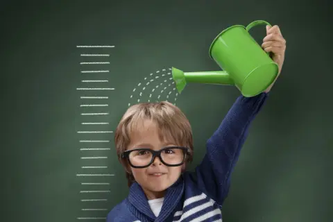 Niño con gafas riega su cabeza con una regadera verde, frente a una tabla de crecimiento que marca el percentil 10.