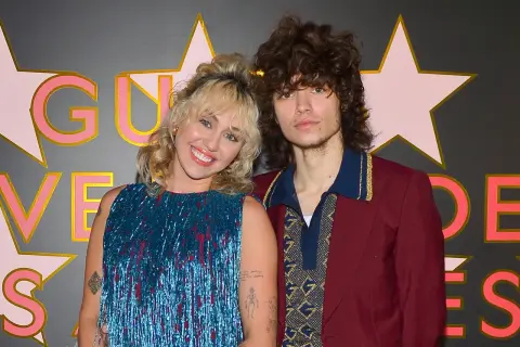 Miley Cyrus y Max Morando posan sonrientes. Ella lleva un vestido azul brillante con flecos, él un traje burdeos con detalles dorados.