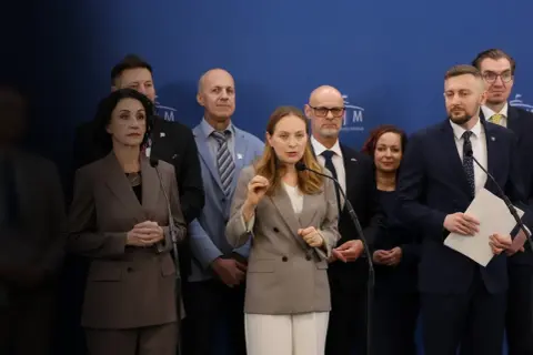 Grupa osób na tle niebieskiej ściany. Kobieta przemawia do mikrofonu, gestykulując. W tle inni mężczyźni i kobiety. Czym Polska 2050 różni się historycznie od nowoczesnej?