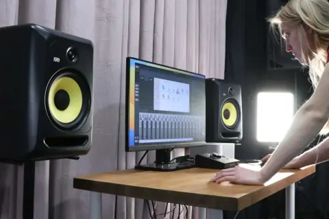 Młoda kobieta pracuje w studiu nagraniowym, otoczona monitorami studyjnymi KRK. Na biurku stoi komputer z programem do produkcji muzyki.