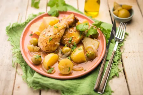 Tajín de pollo con patatas, aceitunas y limón, servido en plato rosa sobre mantel verde.
