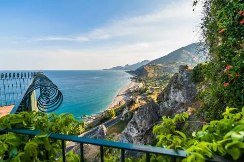 Balkonblick auf Siziliens Küste: Türkises Meer, felsige Hänge & blühende Pflanzen. Perfekt für Sizilien Urlaub Tipps!