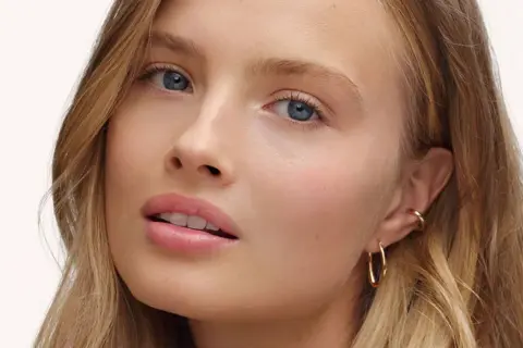 Visage d'une jeune femme aux yeux bleus et aux lèvres rosées, avec un maquillage subtil, style "makeup no makeup". Elle porte des boucles d'oreilles dorées.