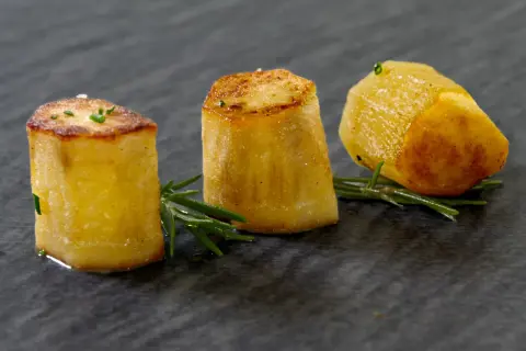 Drei goldbraune Fondantkartoffeln, garniert mit Rosmarin und Schnittlauch, auf dunklem Stein serviert.